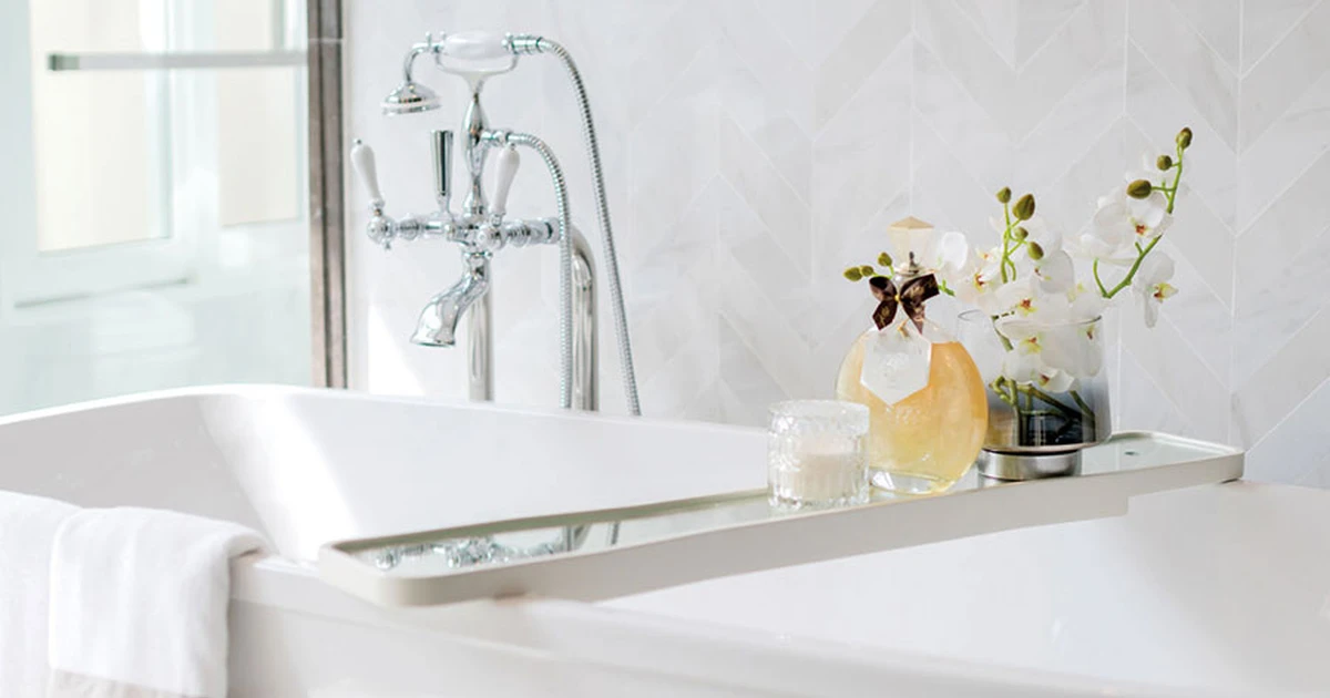 White bathtub with classic chrome mixer taps, a caddy with white orchids, candle & perfume. White herringbone tiled wall.