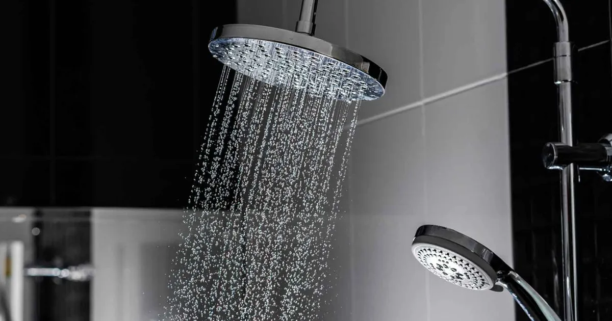 Chrome overhead shower head spraying water, with a handheld shower head visible against black and grey tiles.