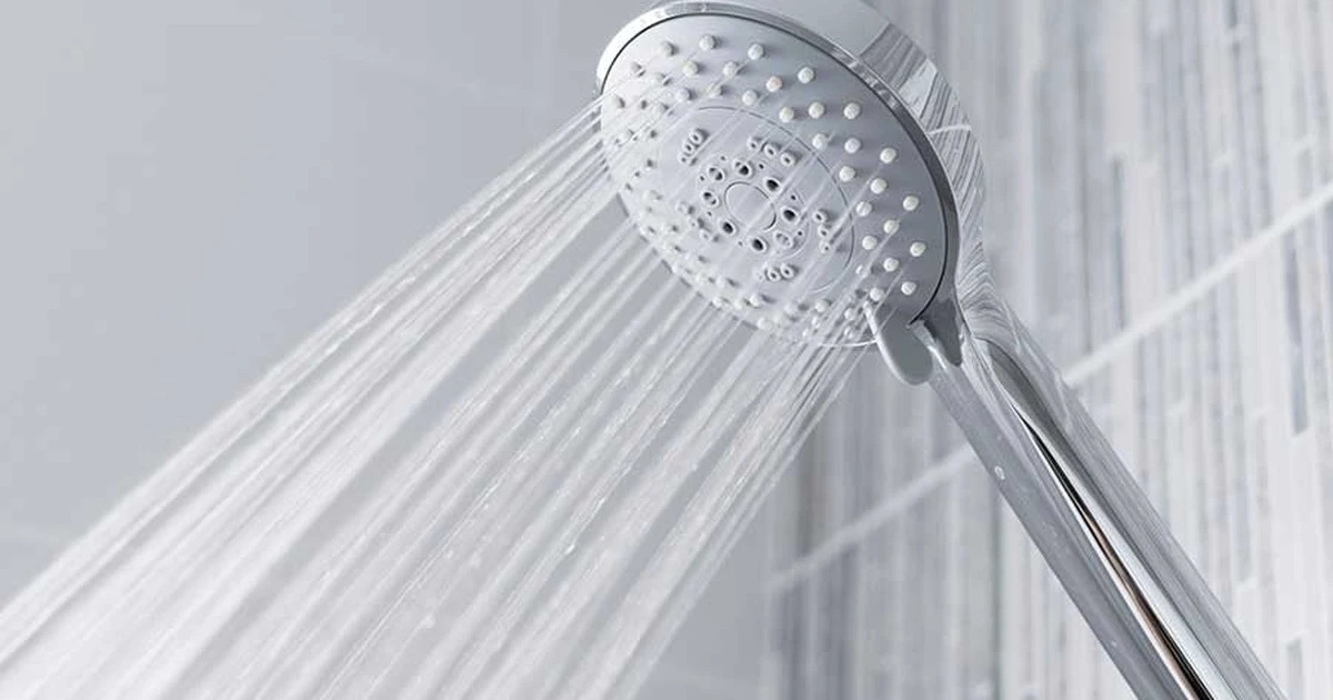 Handheld chrome shower head spraying water against a light grey tiled wall.