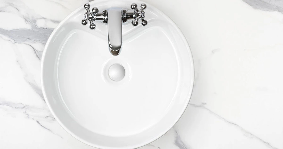 Cross-head chrome mixer tap above a white ceramic basin with plug, on a white marble countertop.